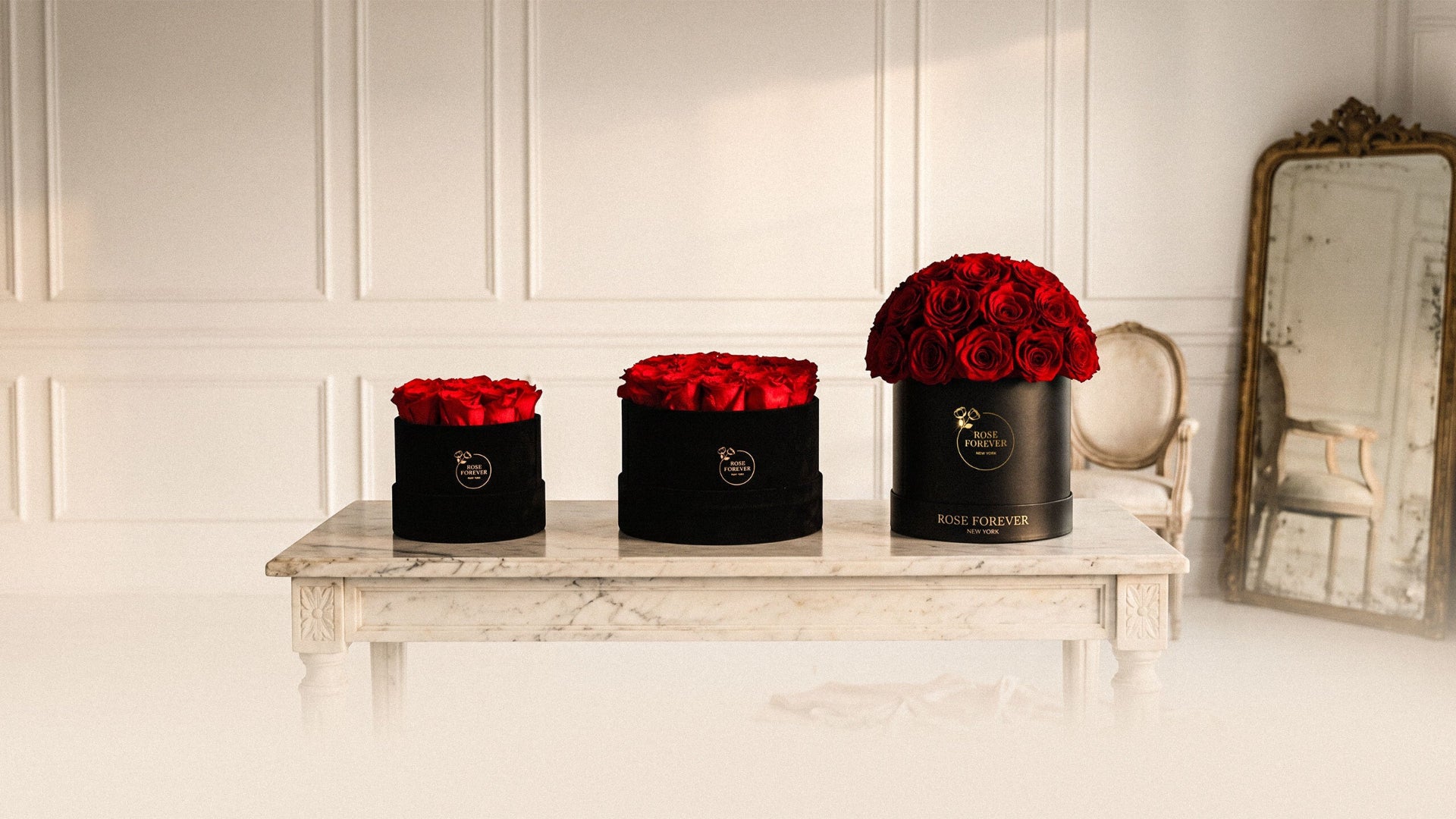 Three black cylindrical flower boxes filled with red roses are displayed on a white marble table in a bright, elegant room with white paneled walls and a vintage mirror.