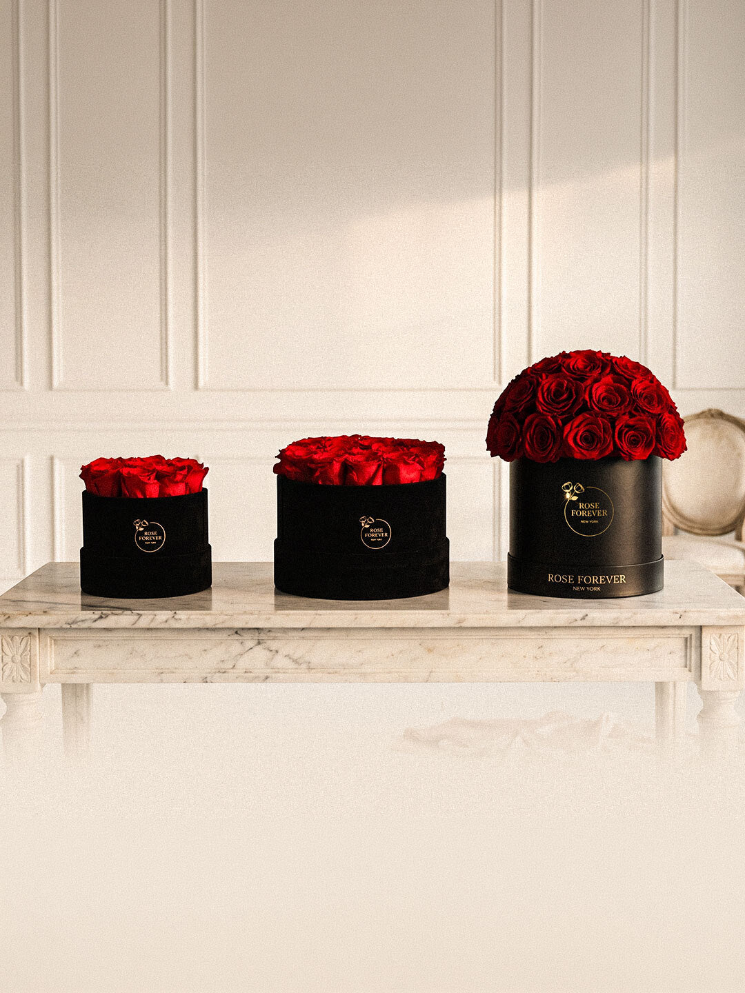 Three black cylindrical flower boxes containing red roses, arranged on a white marble console table against a white paneled wall. The rightmost box is branded ROSE FOREVER.